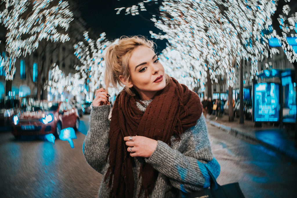 blonde woman with dark red scarf and gray sweater walks along a winter street white christmas lights Lensbaby Featured photos environmental portraits